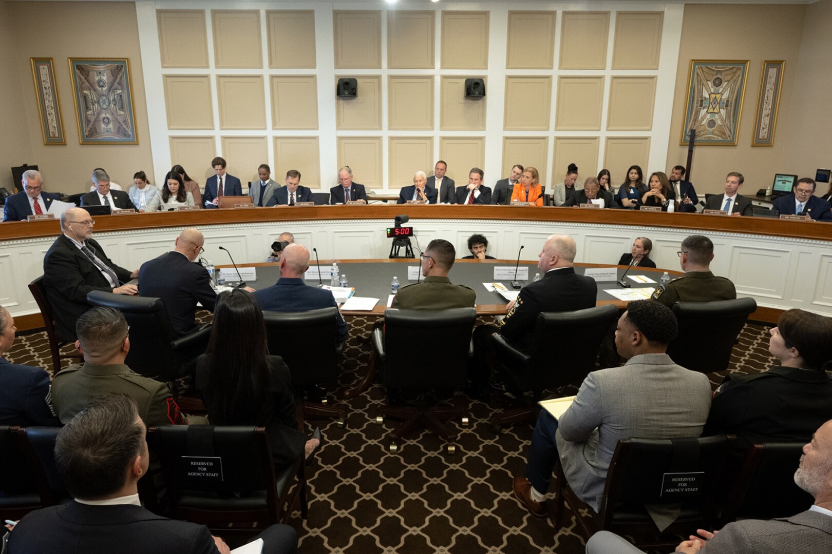 A group of five men in military dress uniforms are sitting at a large table with their backs to the camera while addressing a panel of men and women in civilian attire. The men and women are facing the men in uniform.