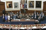 A group of people in business attire and military uniforms sits in a room with flags in the background and portraits hanging on the walls.