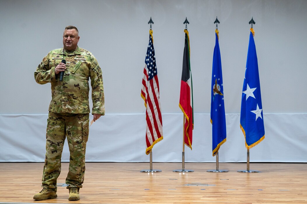 Col. Stouffer stands left of frame holding a microphone as he presents a speech with several flags lined up to his right, including the American flag