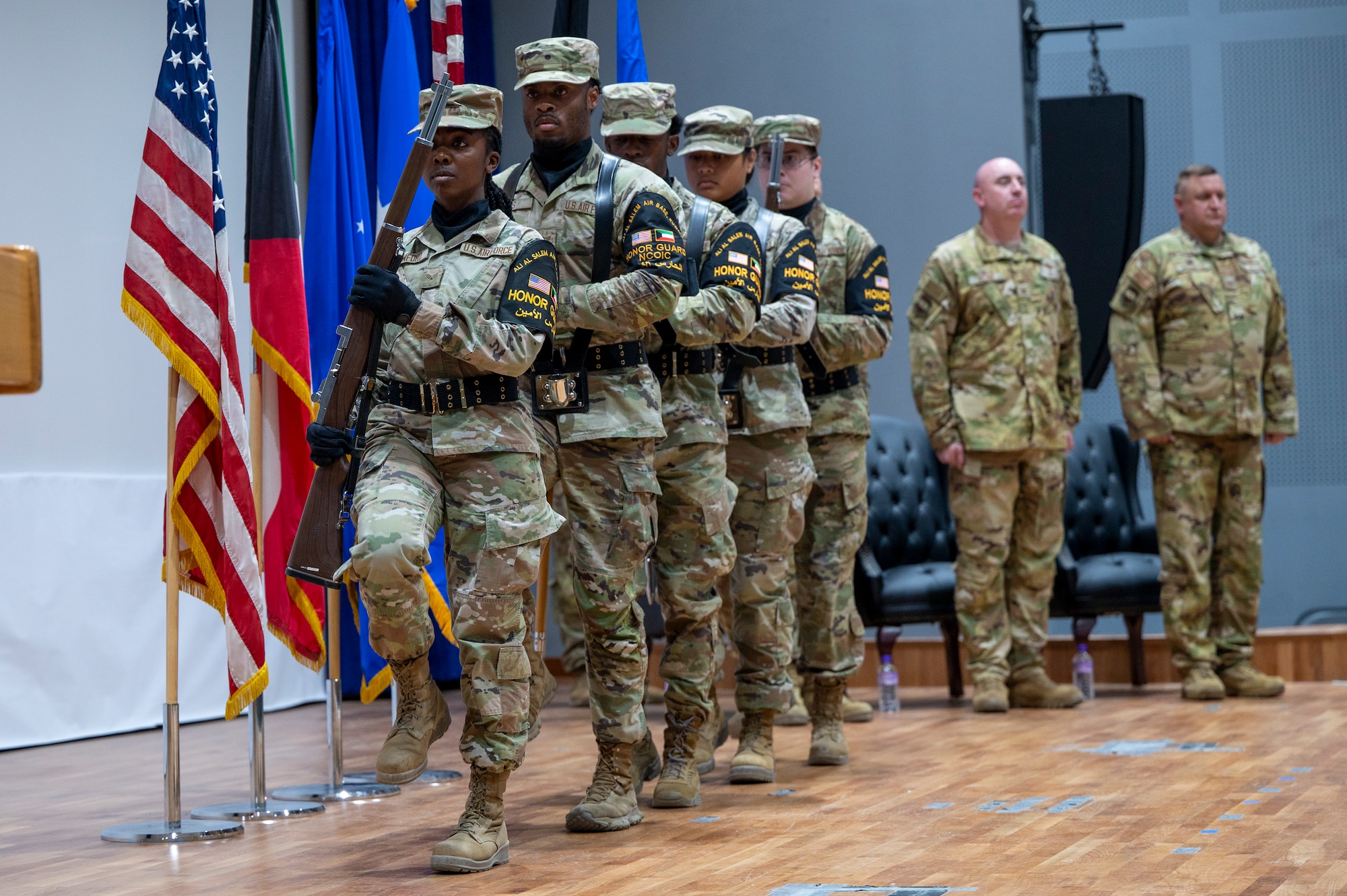 The Honor Guard are lined up leading toward left of frame, marching in unison while the outgoing and incoming commanders stand at attention in the background right of frame