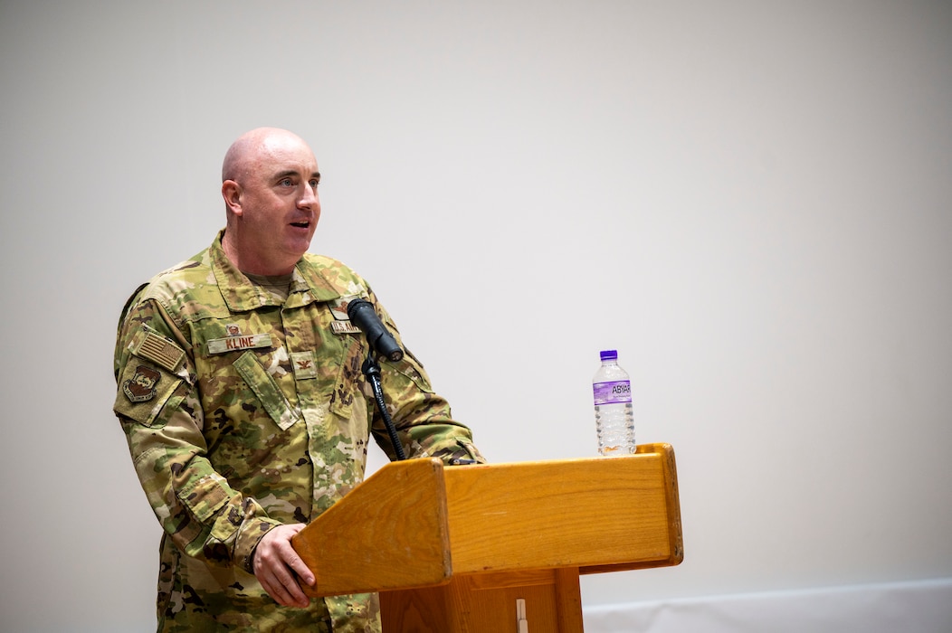 Col. Kline stands left of frame speaking from behind a podium