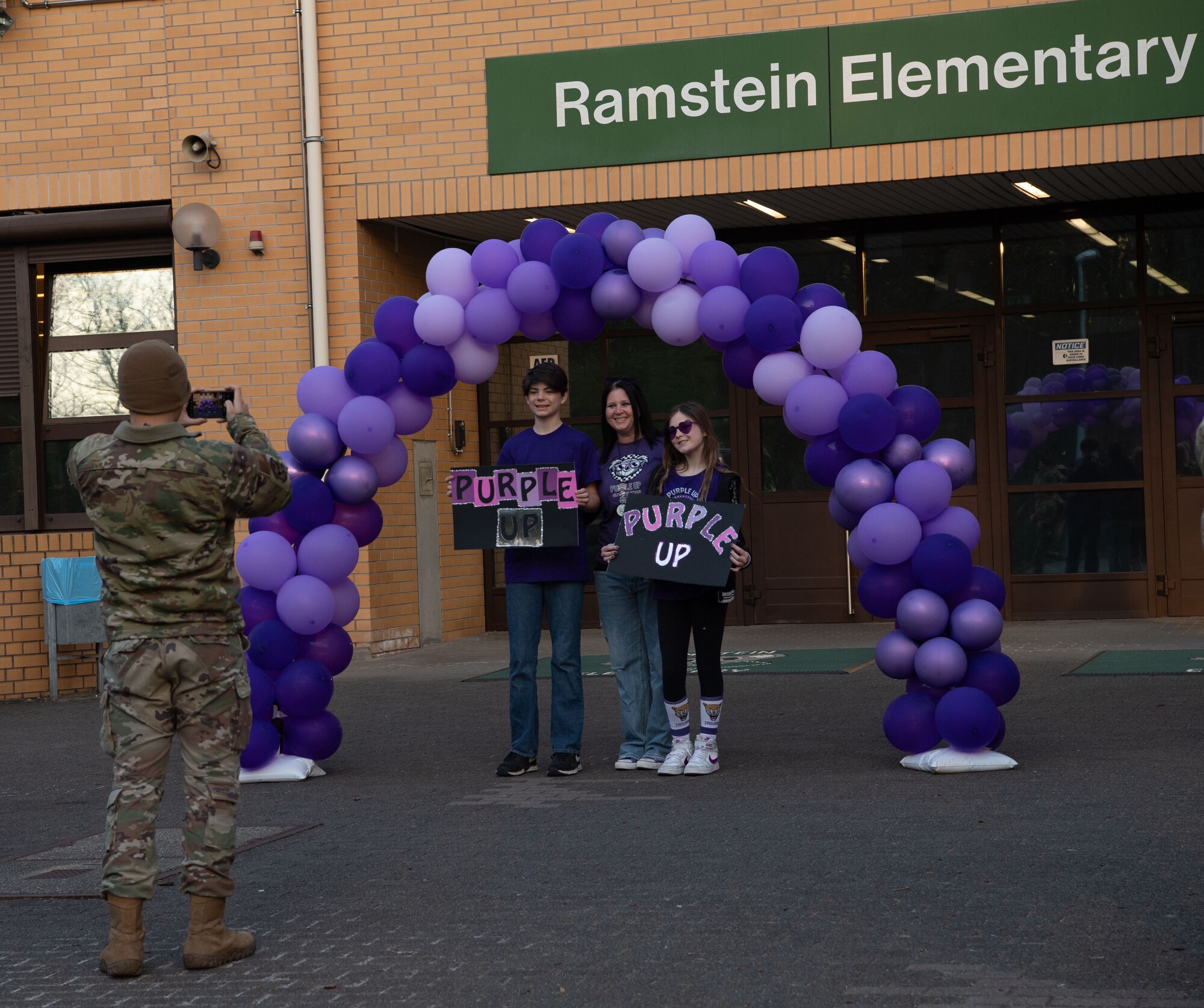 A member of the 86th Security Forces Squadron takes a photo of a mother and child holding their Purple Up! Day signs before students arrive at the Ramstein Elementary School to begin their day at Ramstein Air Base, Germany, April 9, 2025.