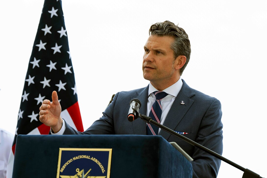 A man in a suit speaks from behind a lectern next to a U.S. flag.