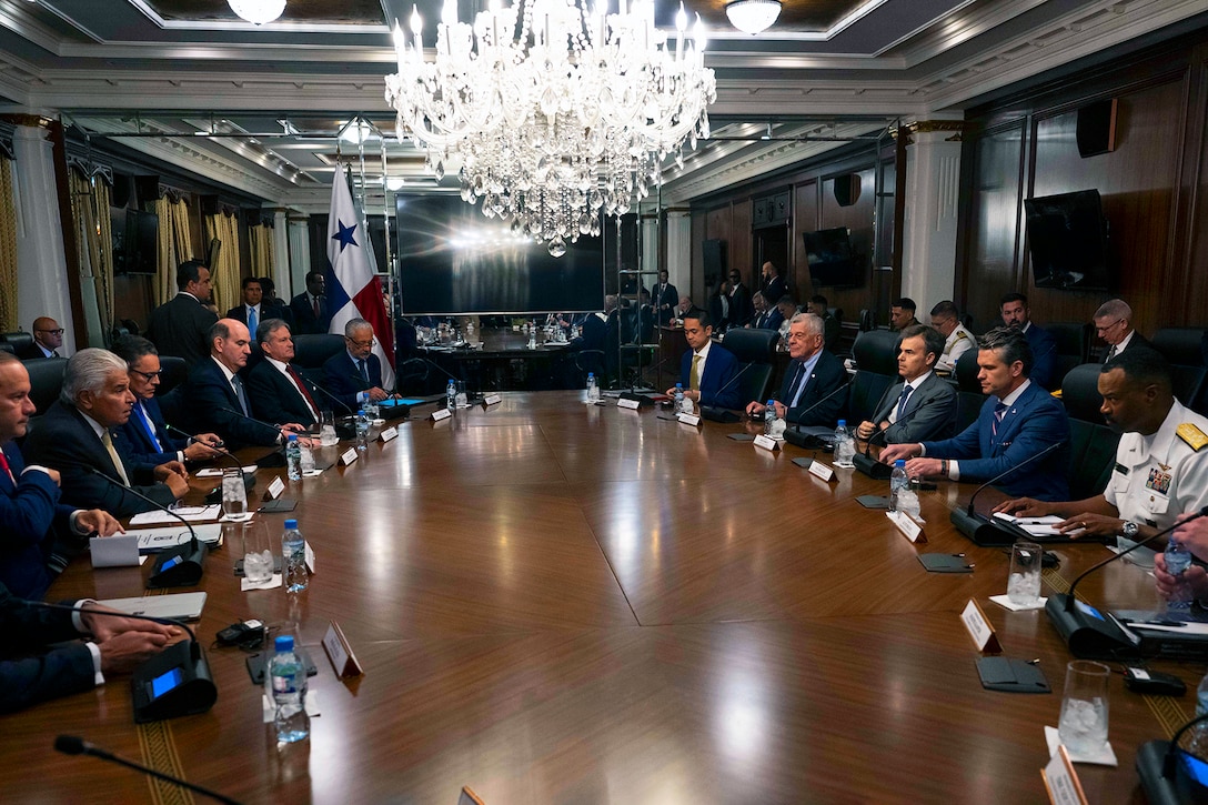 Government officials in suits sit around a table with a large chandelier hanging from the ceiling.