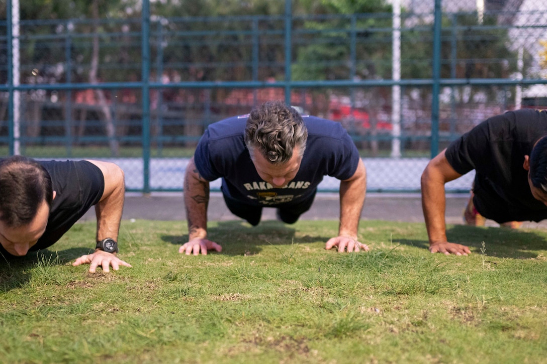 Three men do push ups on the grass in front of a fence outside.