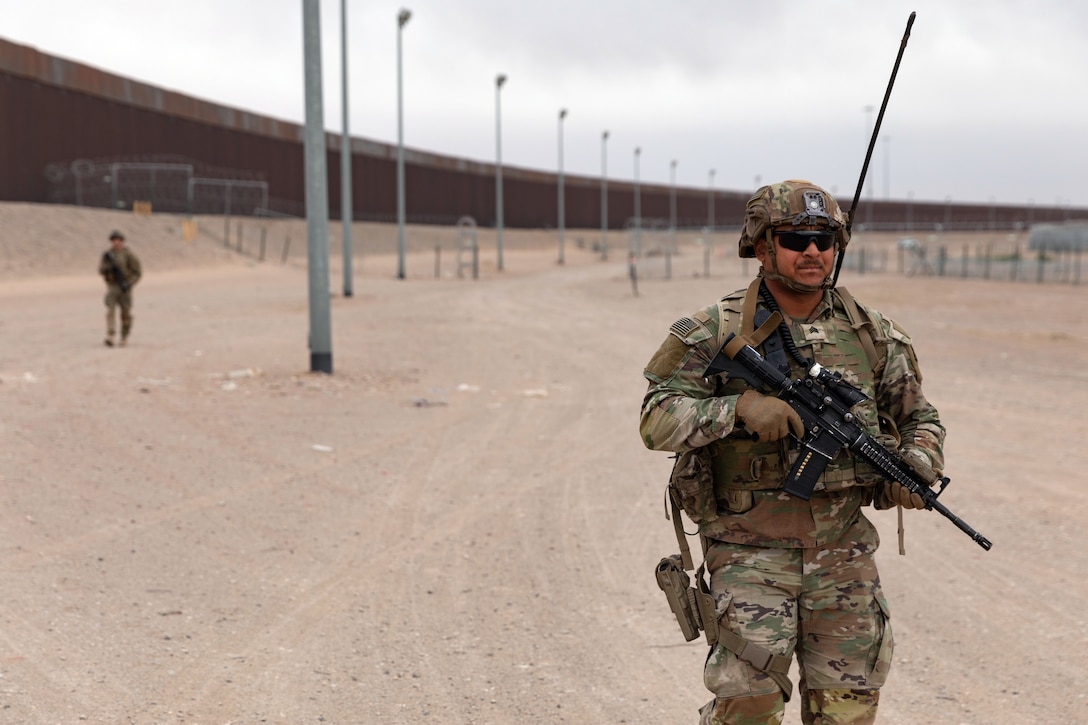 A soldier patrols with a weapon with the southern border wall in the background.
