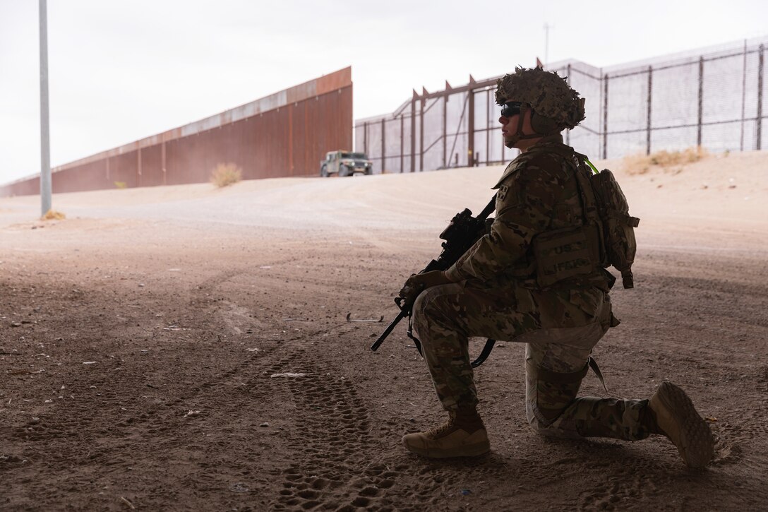 A soldier kneels on a dirt road while a military vehicle and a border wall stands in the background.