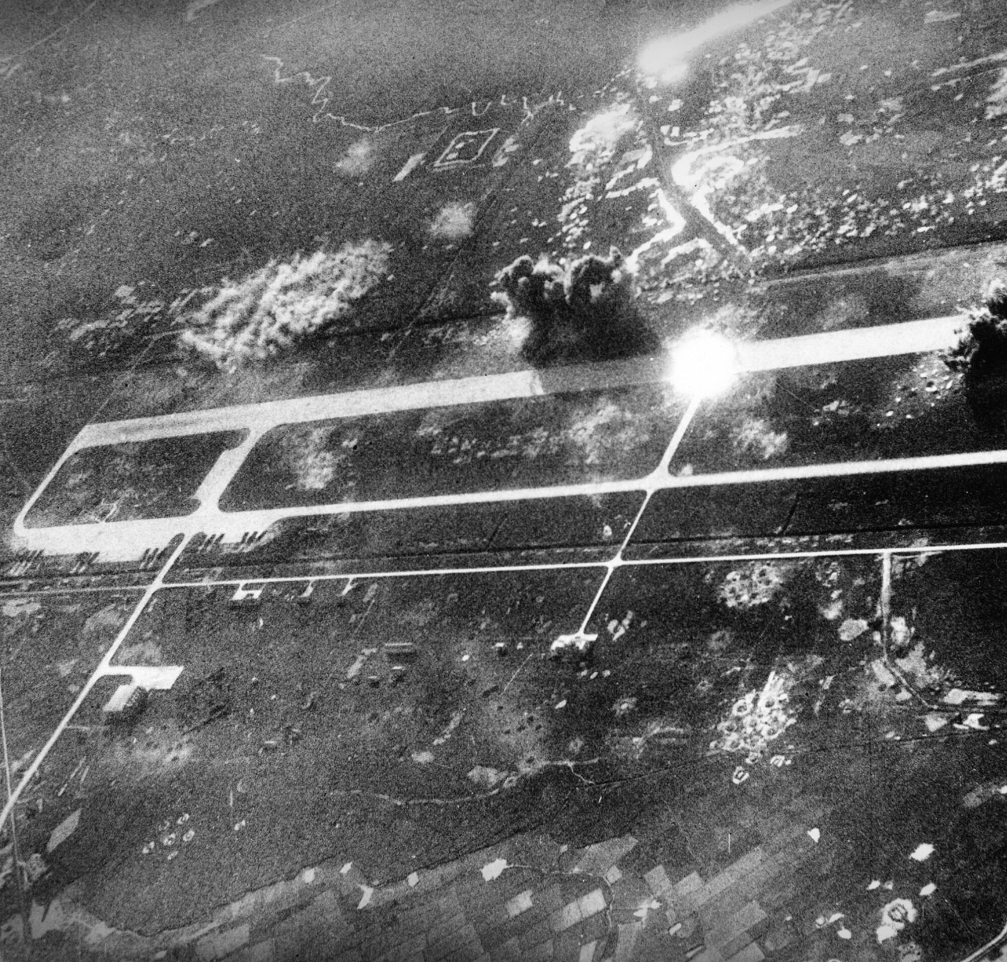 Colonel Edward B. Burdett, commander of the 388th Tactical Fighter Wing, was shot down on November 18, 1967 during a Commando Club radar-assisted raid on Phuc Yen.   The first attacks against Phuc Yen, a MiG-21 Fishbed base north of Hanoi, were authorized in late October, 1967.  The airfield is shown here as USAF bombs hit it during a 1967 attack.  Notice the circular cloud of smoke and dust at the upper left of the picture, probably the detonation of cluster bombs such as CBU-24s dropped by flak-suppression aircraft on an active anti-aircraft artillery site.  Meanwhile, conventional high-explosive bombs, perhaps the commonly used M117 750-lb bombs, explode on or near the runway.  (National Museum of the USAF, via Wikipedia)