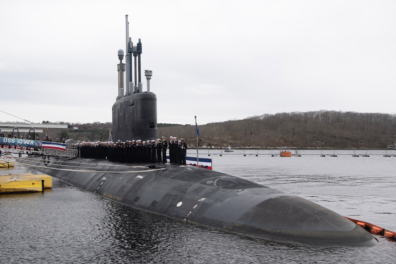 Sailors in uniform stand atop a submarine sitting in the water next to a pier.