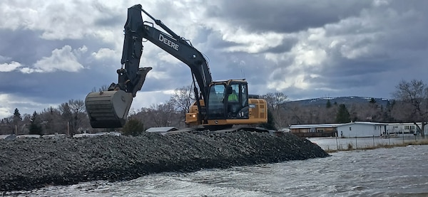 An excavator places rock from a truck on top of already laid rock, water is in the background