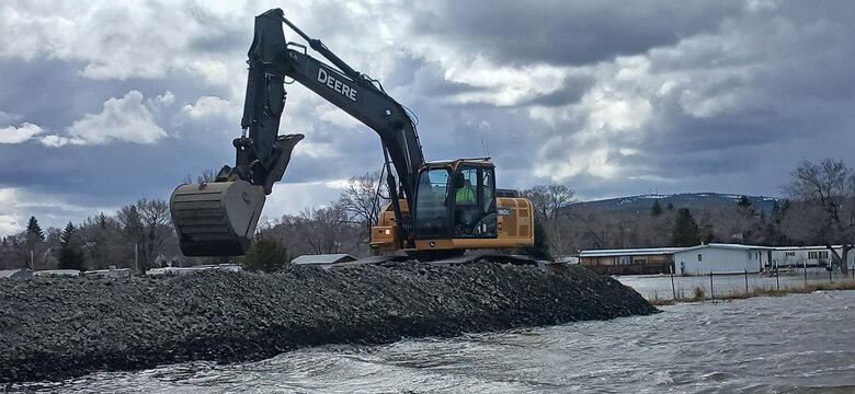 An excavator places rock from a truck on top of already laid rock, water is in the background