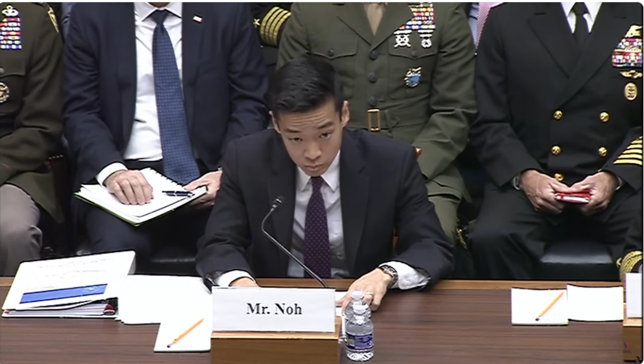 A man in business attire speaks into a microphone while seated behind a table. Three men in military uniforms and one man in business attire sit behind him.
