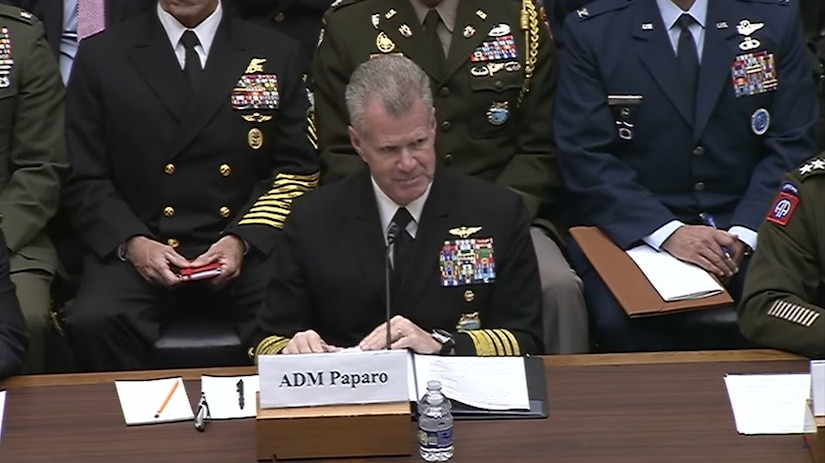 A man in a military uniform speaks into a microphone while seated behind a table. Four men in military uniforms sit behind him.