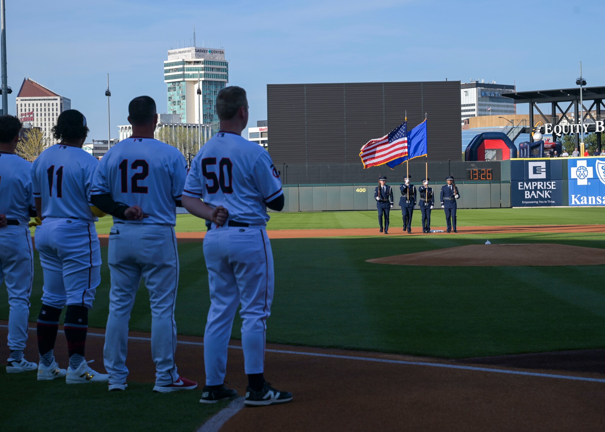 Men is white baseball uniforms stand with their left arm pinned to their back while people in military dress uniforms present the colors.