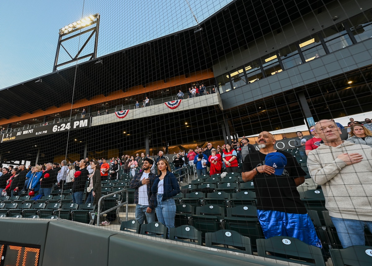People stand in the bleachers of a baseball stadium with their hands over their hearts.