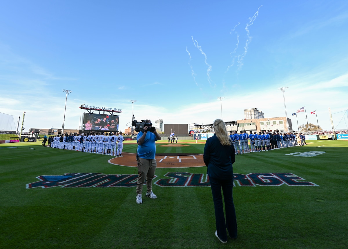 A woman in all black, a man with a large camera on his shoulder and baseball players stand on a baseball field while smoke from fireworks is present in the sky.
