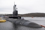 Sailors in uniform stand atop a submarine sitting in the water next to a pier.