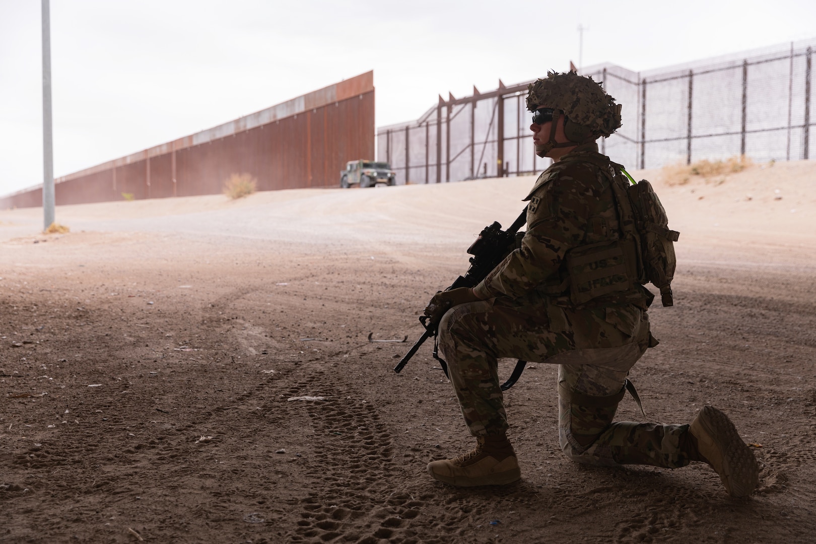 A soldier kneels on a dirt road while a military vehicle and a border wall stands in the background.