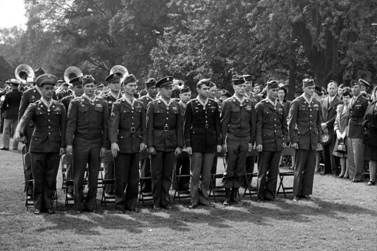 Two rows of men in military uniforms stand at attention as other people watch.