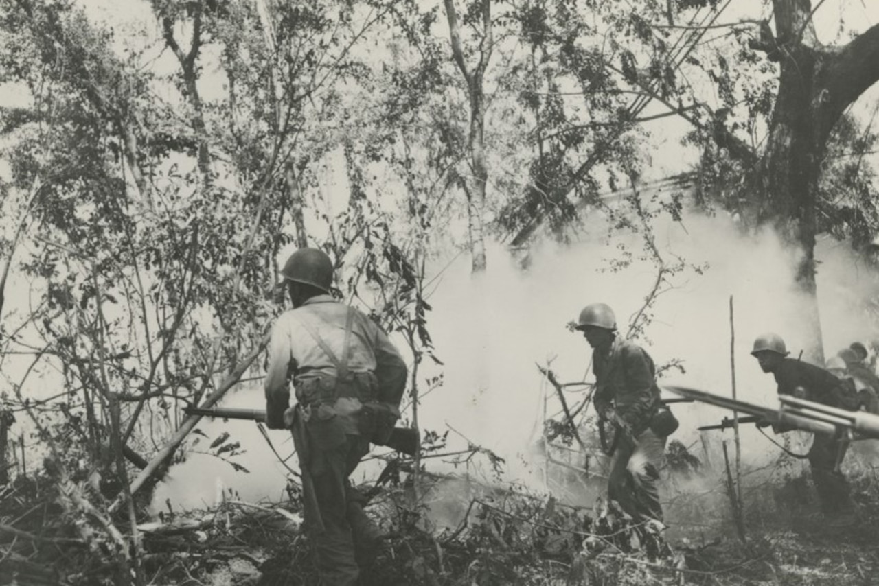 Several men in military uniforms walk into a smoke-filled wooded area.