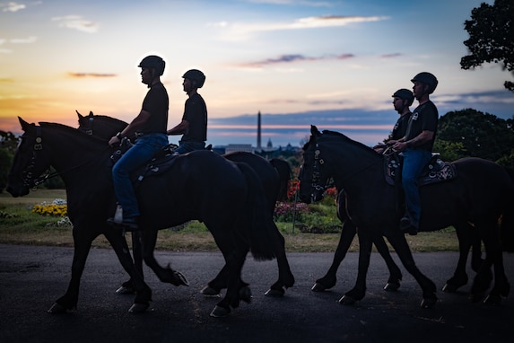4 members of the Army's Caisson Detachment ride on horses for training. It is sunset and the Washington Monument is far in the distance.