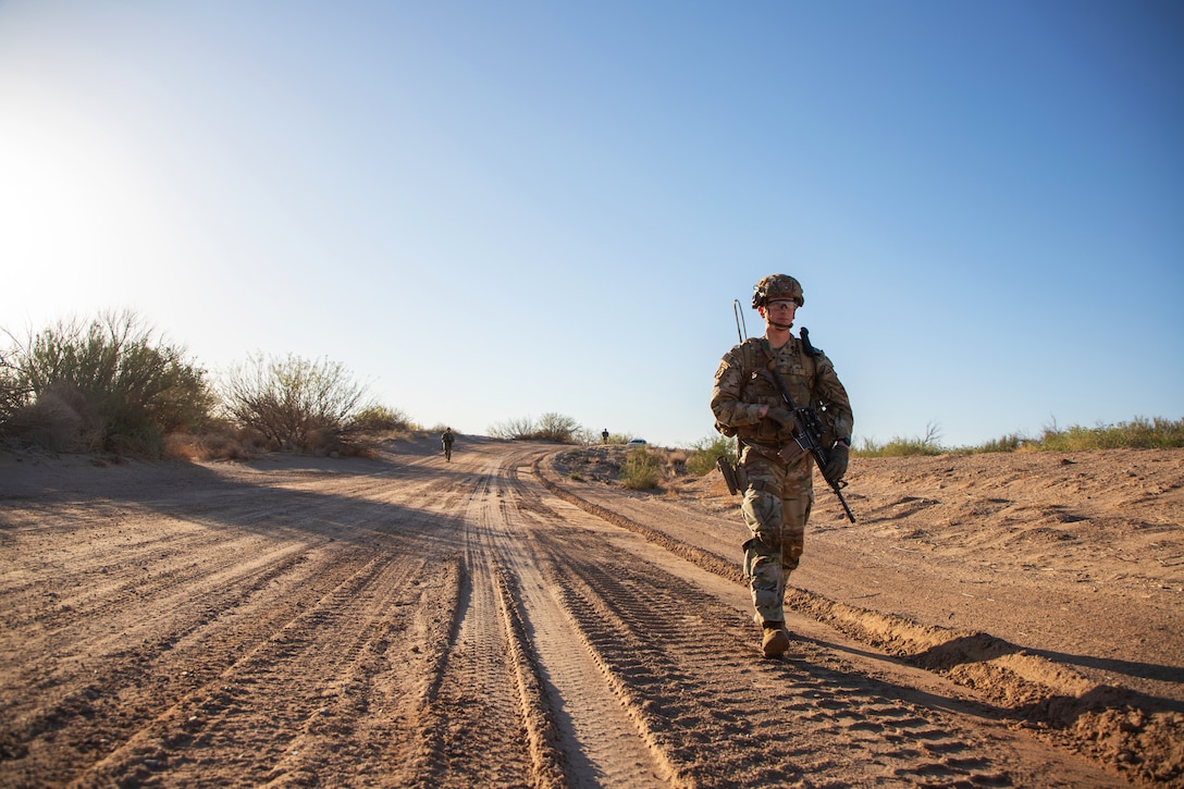 A soldier wearing combat gear and holding a weapon walks along a dirt road in the desert during a sunny day.