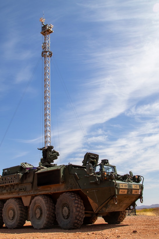 A military vehicle is parked on a desert field next to an observation tower during a sunny day.