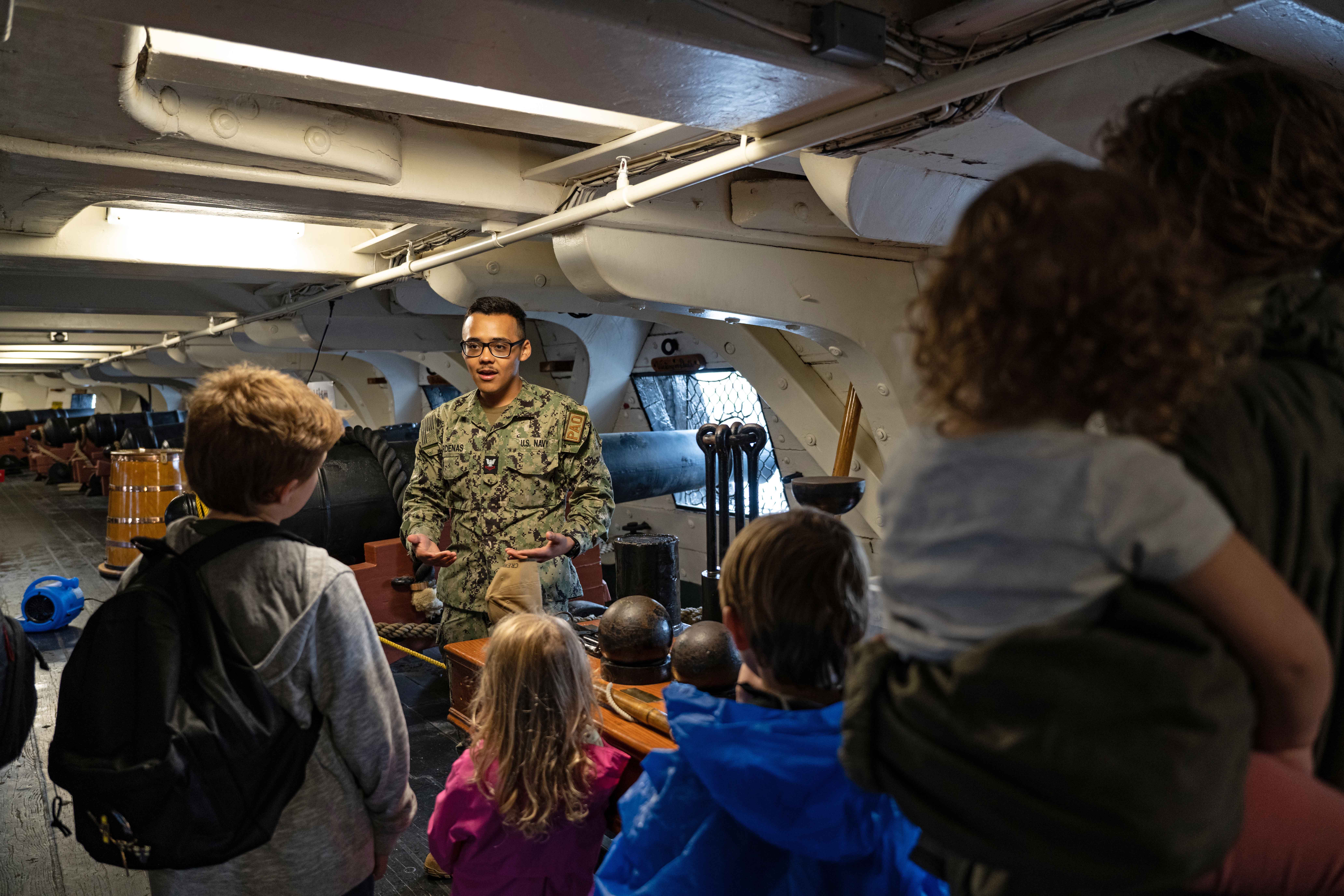 A sailor speaks to a group pf people and children under the deck of an old ship.