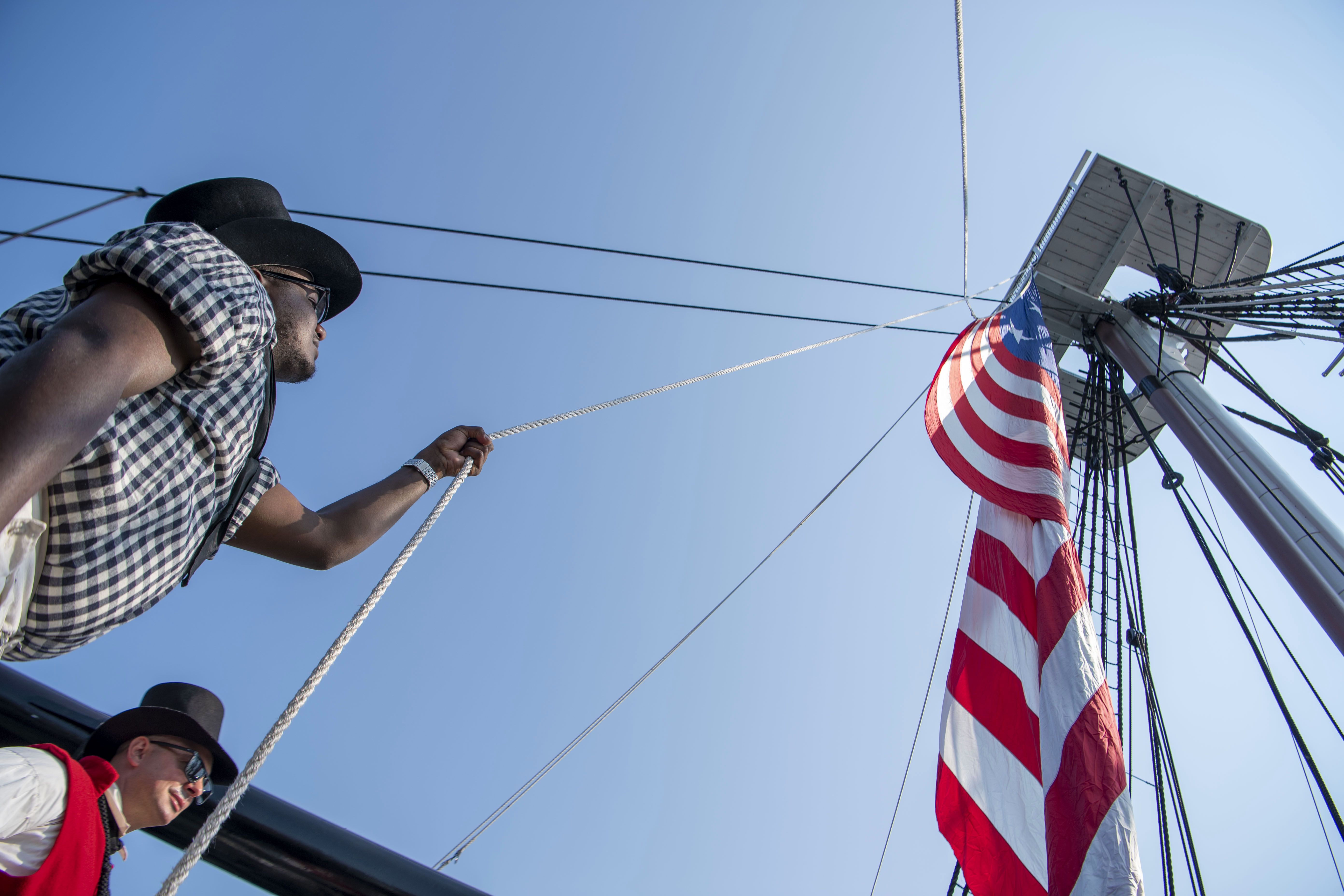 A sailor holds a rope attached to an American flag under the lookout on an old ship, while another sailor watches.