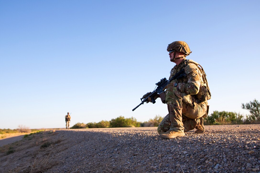 A soldier knees and looks out over a desert as another soldier stands in the distance.