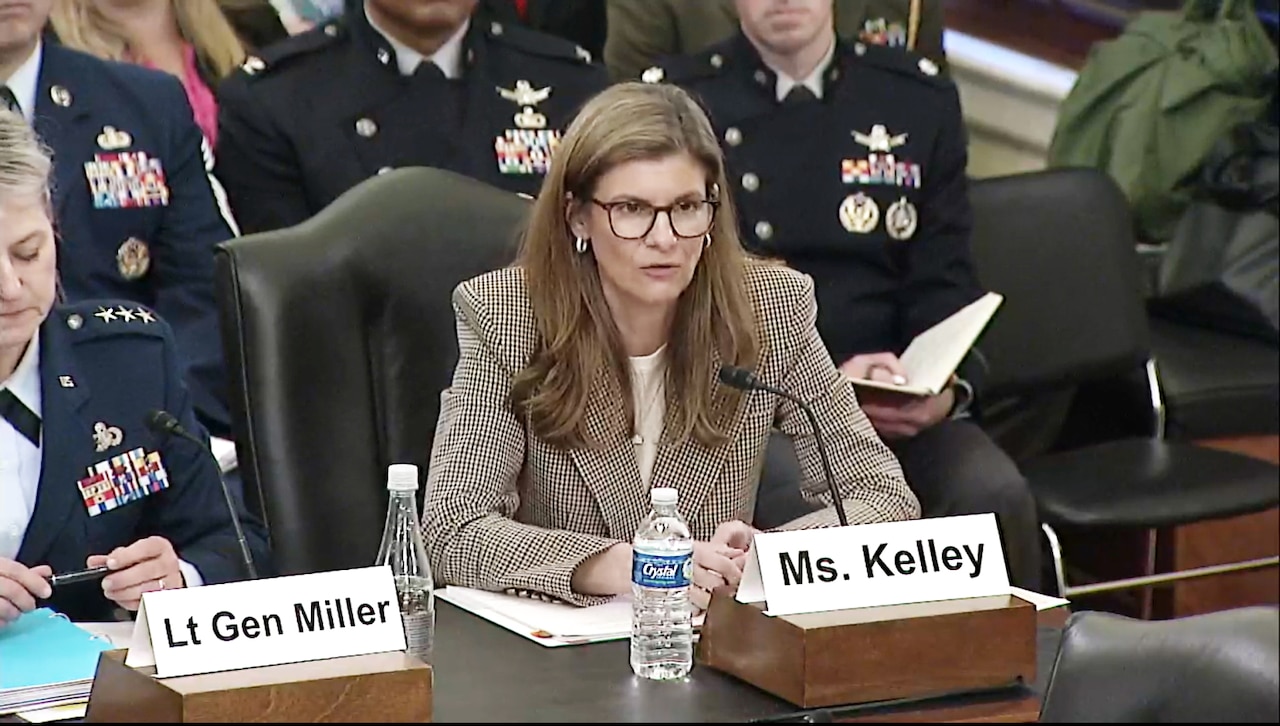 A woman in business attire sits at a wooden table.