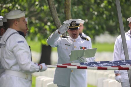 U.S. Navy Sailors wearing white ceremonial uniforms are holding a US flag by its edges in a cemetery as one sailor is holding a book and lifting his right hand into the air while speaking during a funeral.