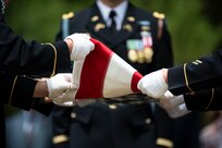Army soldiers wearing dark ceremonial uniforms and white gloves are folding a US flag during a funeral.