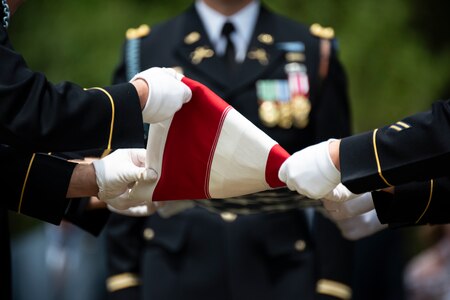 Army soldiers wearing dark ceremonial uniforms and white gloves are folding a US flag during a funeral.