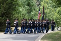 Army Soldiers in dark ceremonial uniforms and carrying rifles in front of their chests are marching down a street in Arlington National Cemetery.
