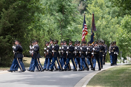 Army Soldiers in dark ceremonial uniforms and carrying rifles in front of their chests are marching down a street in Arlington National Cemetery.