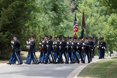Army Soldiers in dark ceremonial uniforms and carrying rifles in front of their chests are marching down a street in Arlington National Cemetery.