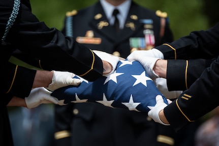 Army Soldiers wearing dark ceremonial uniforms with white gloves are folding a US flag.