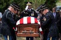 Army Soldiers in dark ceremonial uniforms a lowering a US flag-draped casket down at gravesite while another Soldier salutes.