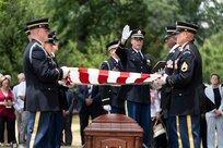 Army soldiers are holding the edges of a US flag over a brown wooden casket at an outdoor military funeral as a chaplain raises his right hand and gestures as he prays over the casket.