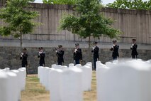 Seven Army Soldiers dressed in dark ceremonial uniforms are holding rifles and preparing to fire them as a 21-gun salute in a cemetery.