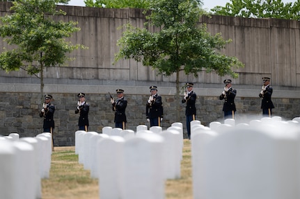 Seven Army Soldiers dressed in dark ceremonial uniforms are holding rifles and preparing to fire them as a 21-gun salute in a cemetery.