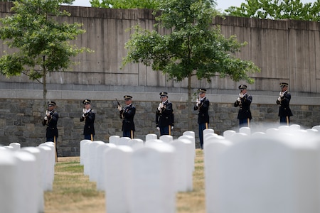 Seven Army Soldiers dressed in dark ceremonial uniforms are holding rifles and preparing to fire them as a 21-gun salute in a cemetery.