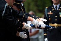 Army soldiers wearing dark ceremonial uniforms are passing a folded US flag during a funeral.