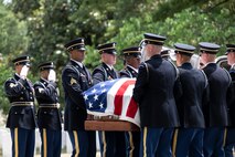 Army Soldiers dressed in dark ceremonial uniforms are carrying a US flag-draped casket while other soldiers nearby are saluting.
