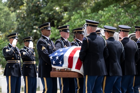 Army Soldiers dressed in dark ceremonial uniforms are carrying a US flag-draped casket while other soldiers nearby are saluting.