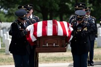 Army Soldiers dressed in dark ceremonial uniforms are looking down while carrying a brown, wooden casket that has a US flag draped across it.