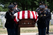 Army Soldiers dressed in dark ceremonial uniforms are looking down while carrying a brown, wooden casket that has a US flag draped across it.