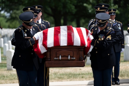 Army Soldiers dressed in dark ceremonial uniforms are looking down while carrying a brown, wooden casket that has a US flag draped across it.