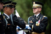 An Army Soldier dressed in dark ceremonial uniform is holding a folded US flag with both hands while another Soldier is saluting the flag.